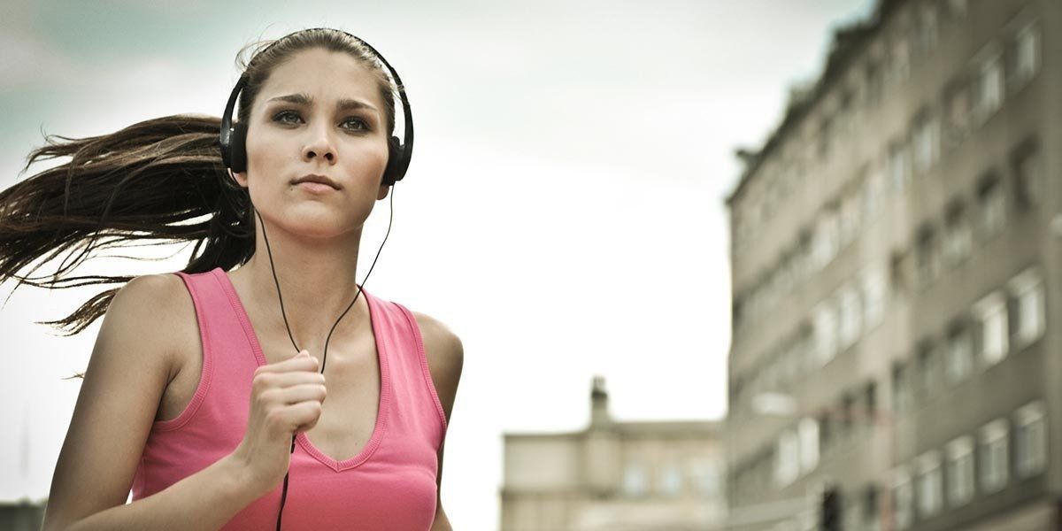 Woman jogging outdoors with earbuds, wearing a pink tank top, city buildings in the background