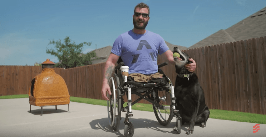 Man in wheelchair outdoors with a black dog, beside an orange smoker and wooden fence under a blue sky