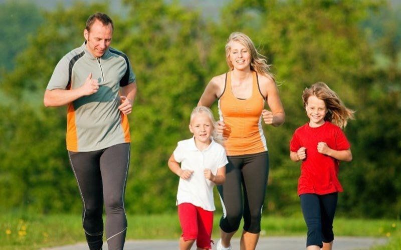 Family jogging together on a sunny outdoor path, wearing athletic clothes, smiling and exercising