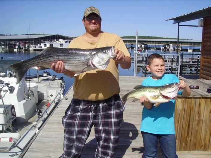 Two people on a dock holding large fish, with boats and a marina in the background.
