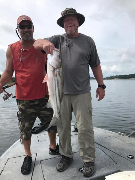 Two anglers standing on a boat deck, holding a large fish on a calm lake.