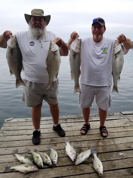 Two anglers on a dock holding several fish, with more fish laid out at their feet.