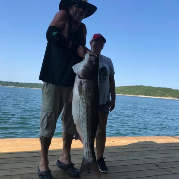Two people on a dock holding a large striped fish by the water under a blue sky