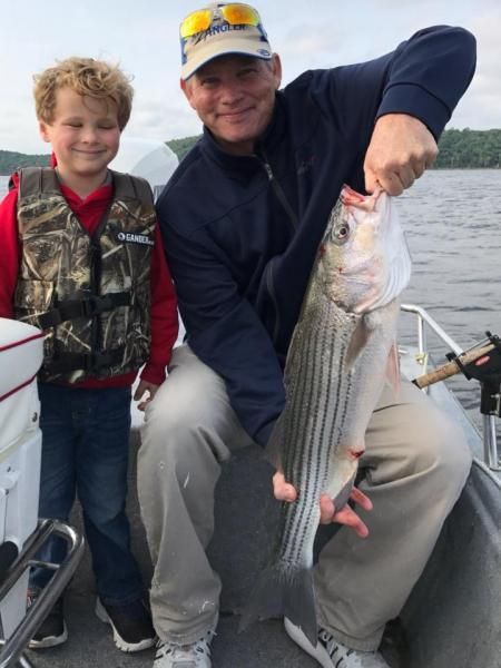 Two people on a boat holding a large striped fish, with calm water in the background.