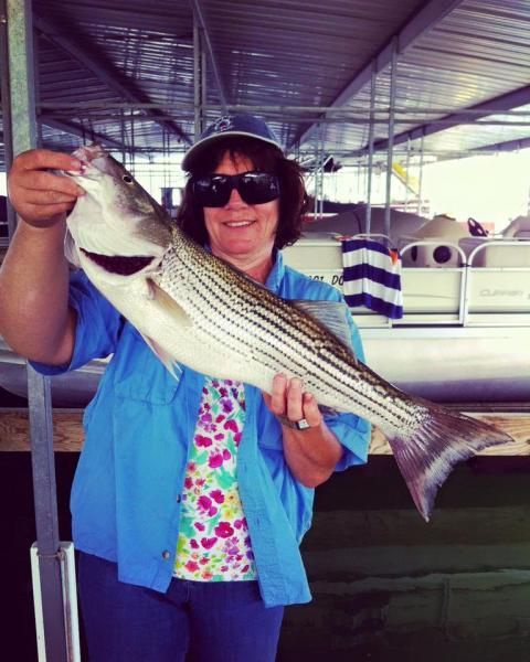 Person holding a large striped fish in a covered marina, smiling and wearing sunglasses.