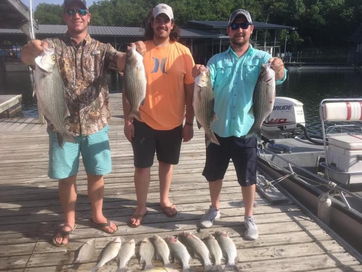 Three anglers on a dock hold up fish with a row of catches laid out at their feet.