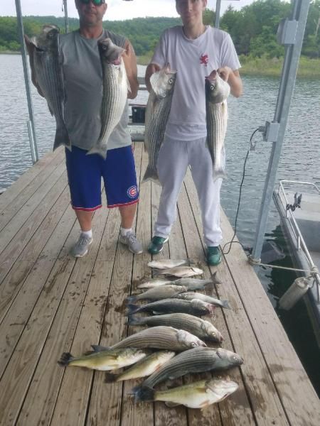 Two people on a dock holding large fish, with a pile of catch at their feet by the water.