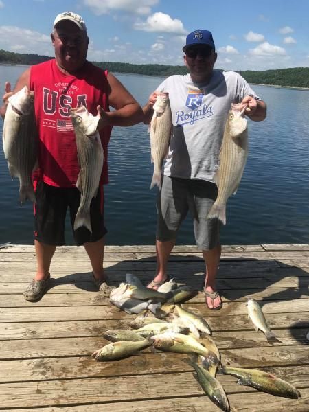 Two people on a dock holding large fish, with several fish laid out at their feet by a lake.