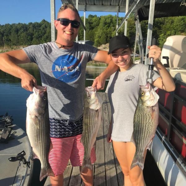 Two people holding large striped fish on a boat by the water, smiling in the sun.
