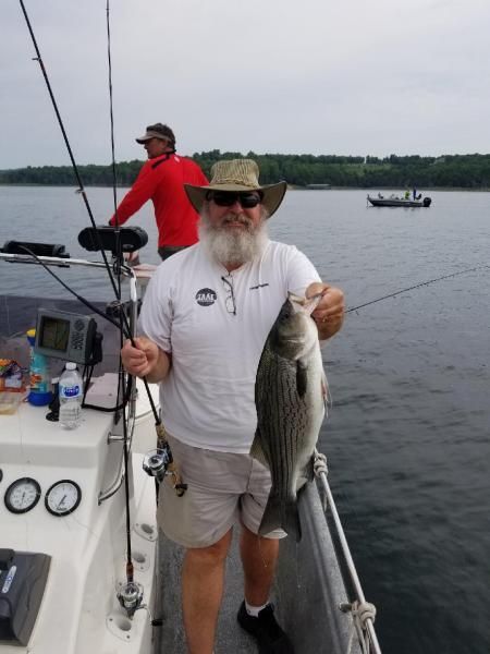 Man on a boat holds a striped fish, with another angler and calm lake in the background.
