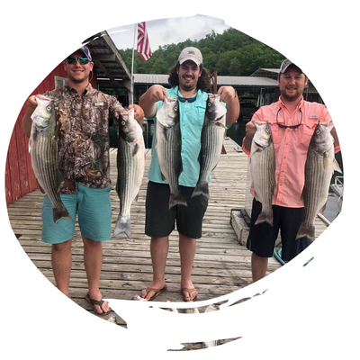 Three anglers on a dock, each holding up large fish, with boats and an American flag in the background