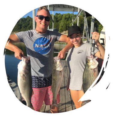 Two people holding large fish on a dock, smiling beside a boat in bright daylight.
