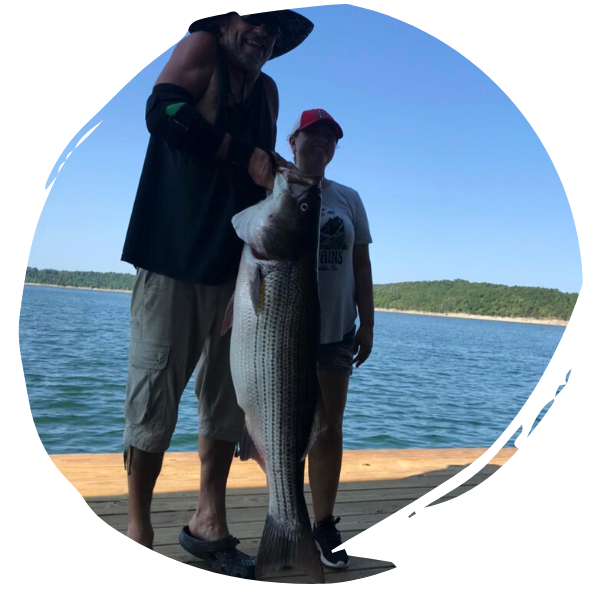 Two people standing on a dock, holding a large fish by a lake under a blue sky.