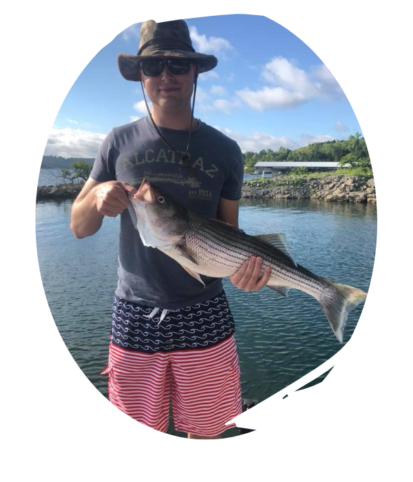 Person holding a large striped fish beside a calm body of water under a partly cloudy sky