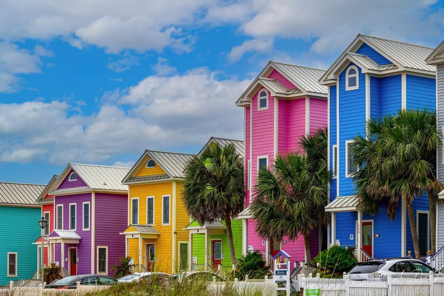 Colorful beachfront houses, ranging in hues of teal, purple, yellow, and blue, under a blue sky.