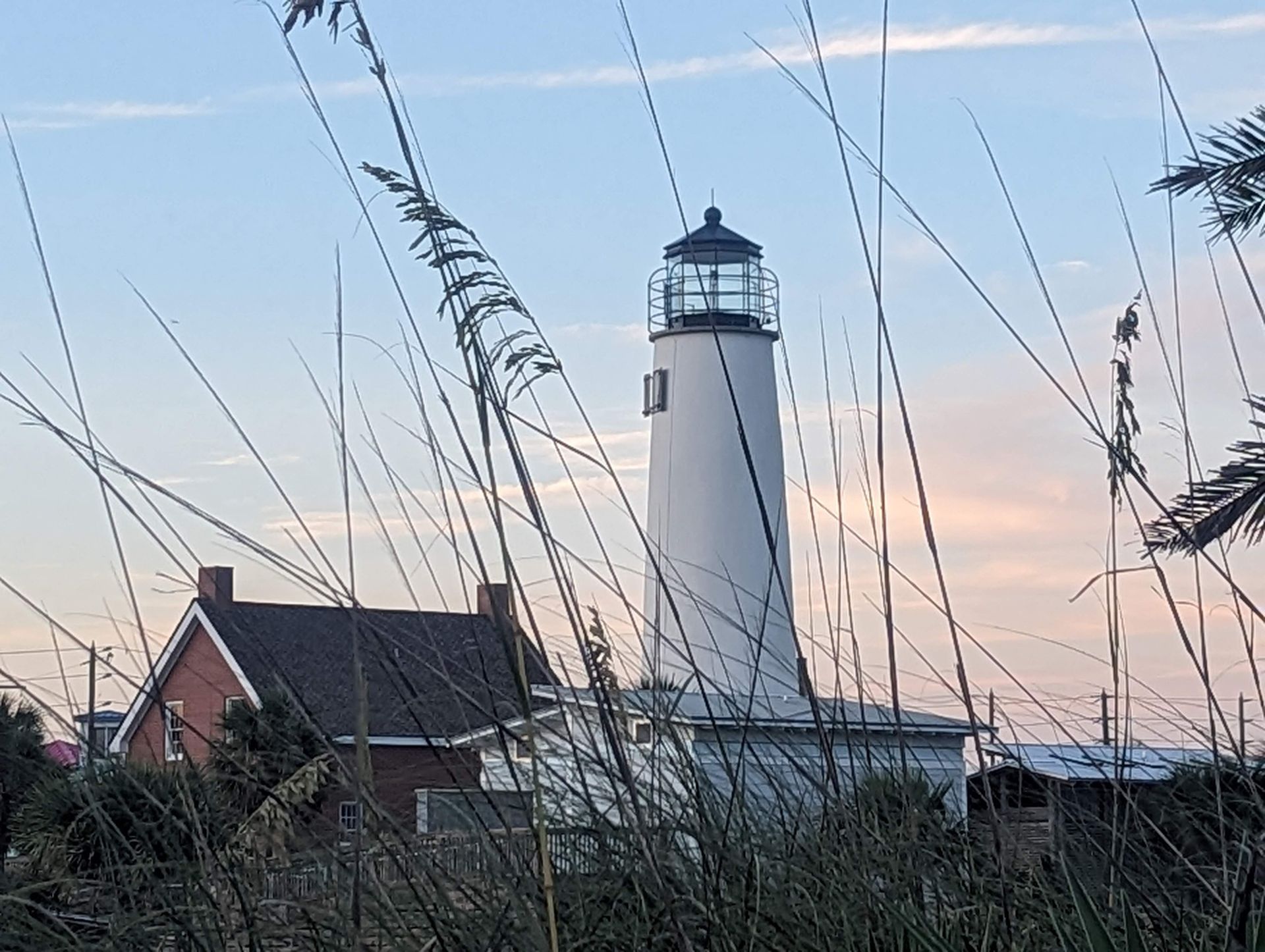 White lighthouse with attached red brick building against a soft sunset sky, seen through beach grasses.