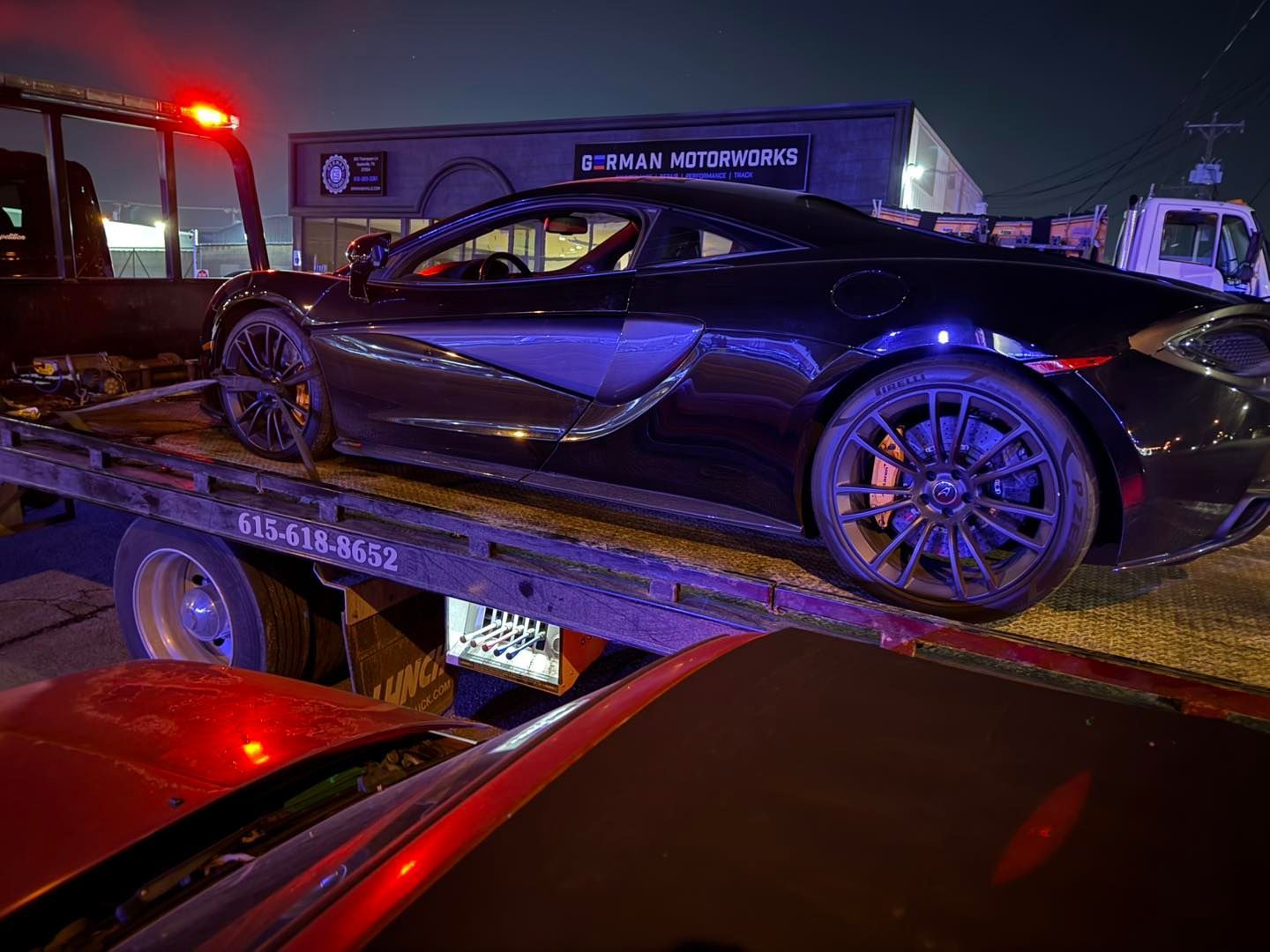 Black McLaren sports car being towed at night. Tow truck in front of a building with a lit sign.