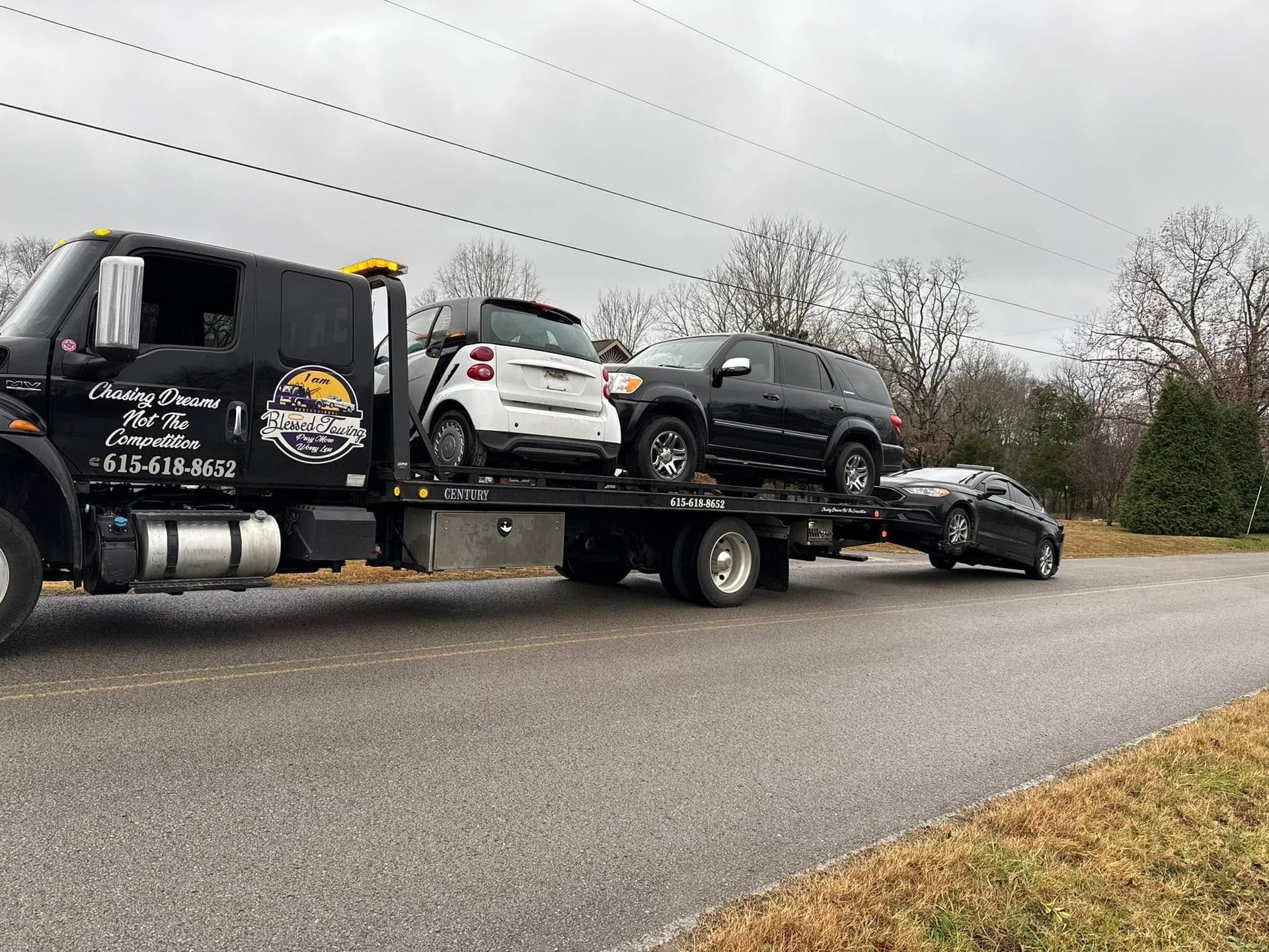 Tow truck carrying three vehicles on a road. Black truck with white, black, and silver cars. Overcast sky.
