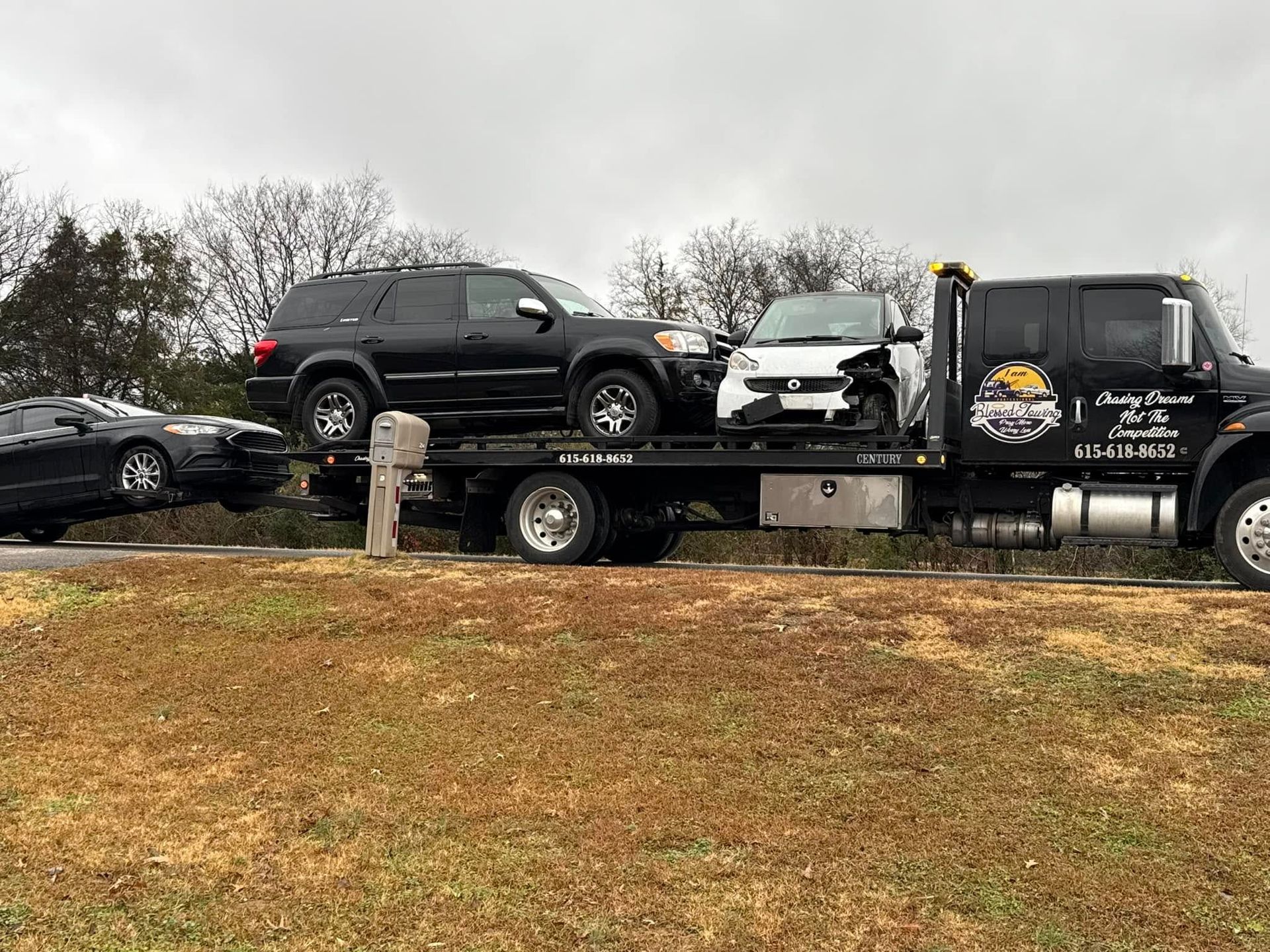 Tow truck carrying two vehicles on a highway; a black SUV and a damaged white car. Overcast sky.