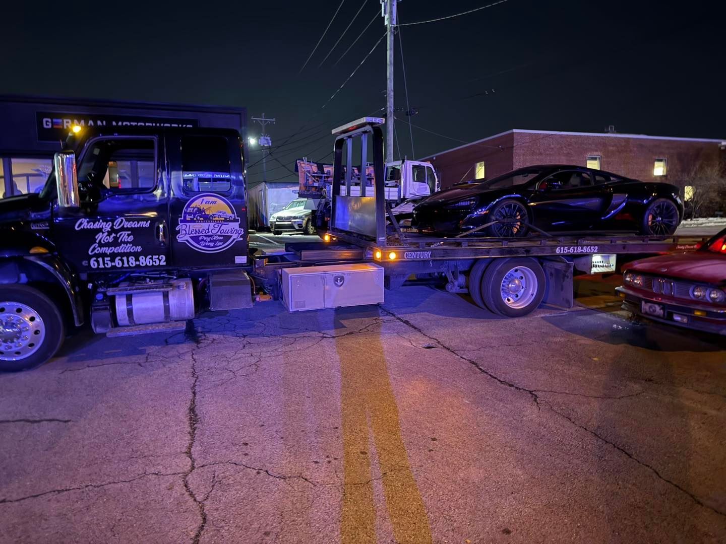 A black sports car being towed on a flatbed truck at night on a street with yellow lines.