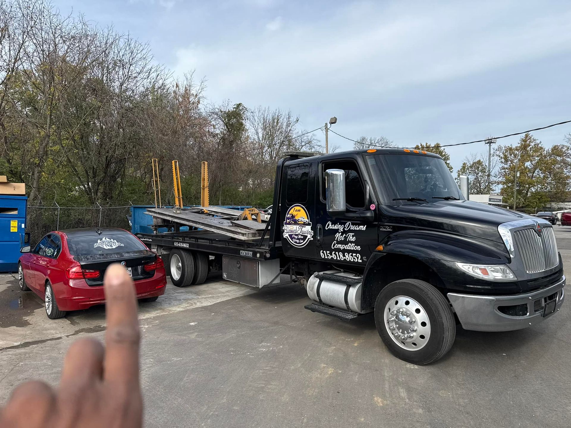 Tow truck loading a red car on a sunny day. Finger pointing towards the car. Black truck with logo.