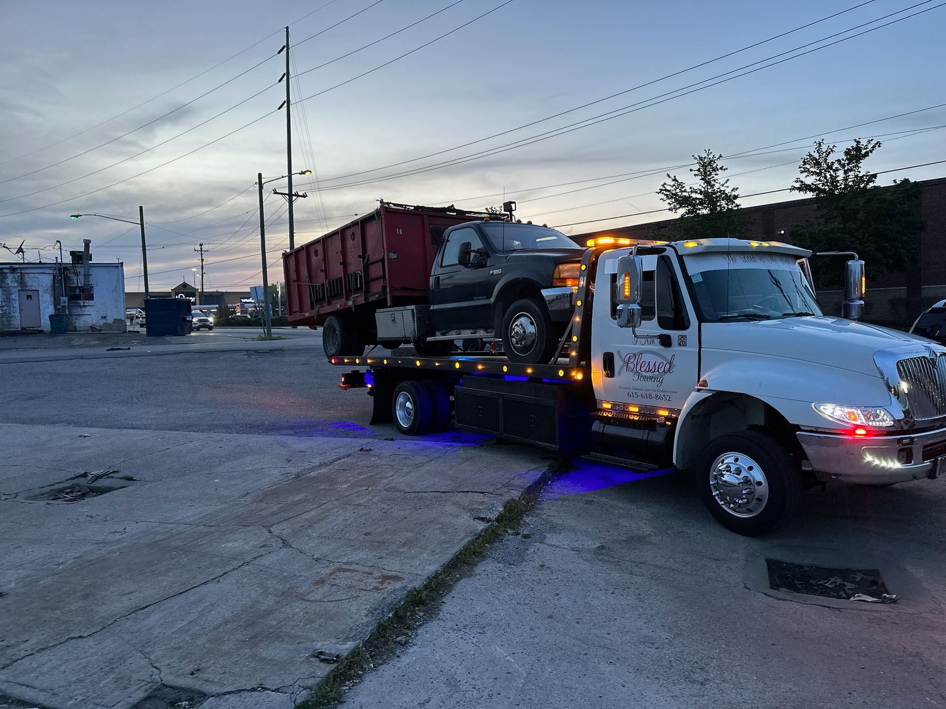 Tow truck carrying a black vehicle on its flatbed at dusk. Red dumpster visible.