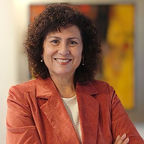 Una mujer con cabello castaño rizado sonríe con un blazer de color óxido; arte borroso en el fondo.