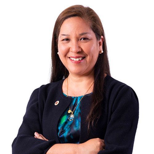 Mujer sonriente con los brazos cruzados, vistiendo un blazer y un collar colgante.