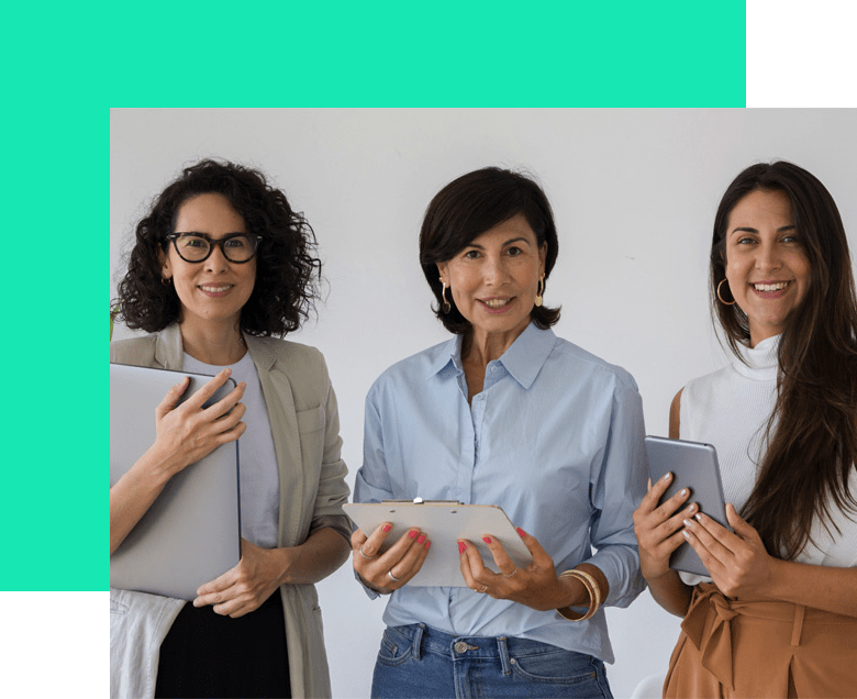 Tres mujeres sonrientes sosteniendo una computadora portátil, un portapapeles y una tableta, de pie frente a una pared iluminada.