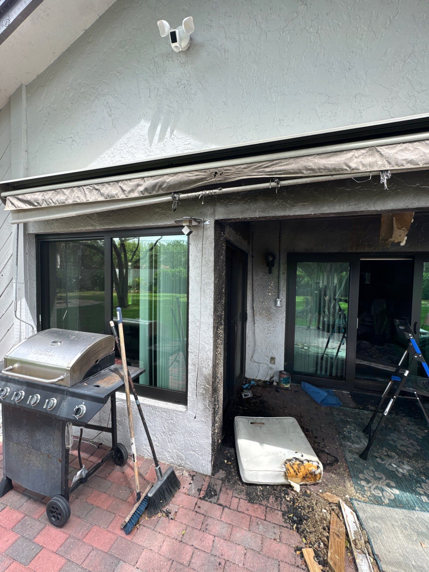 Damaged patio sliding door and exterior wall with debris, grill, and tools on a brick patio