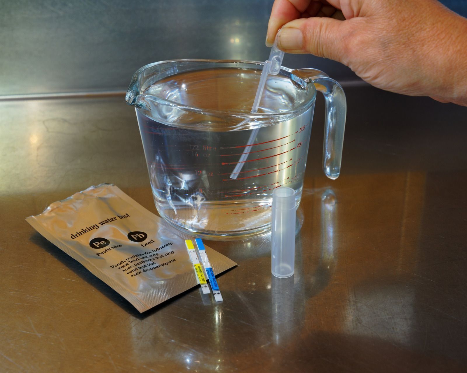 Hand pouring liquid into a clear measuring cup on a stainless steel counter beside a small packet and dropper strip