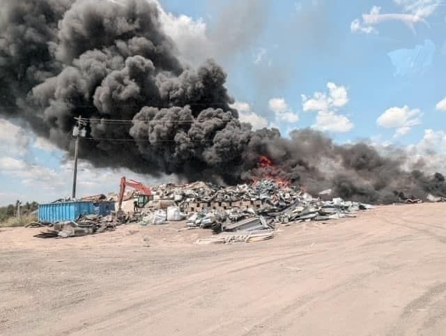 Thick black smoke rises from a large pile of burning scrap in a dusty open yard