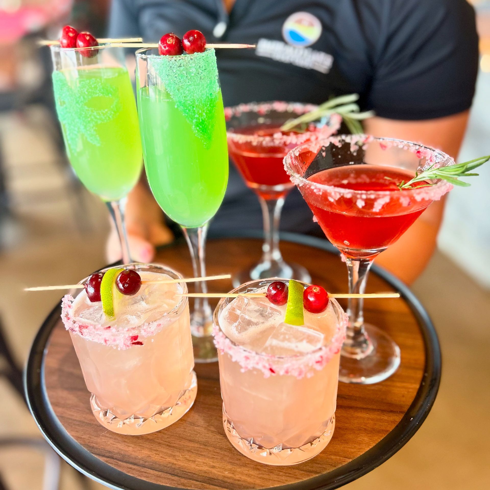 A server holds a tray featuring two green champagne-style drinks, two red cocktails, and two light pink drinks on ice.