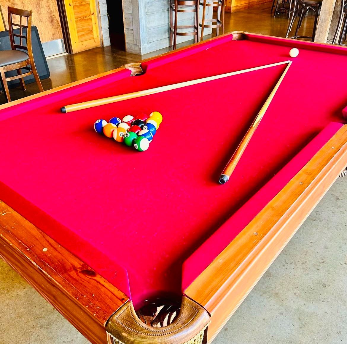 A pool table with red felt, a rack of billiard balls, and two pool cues positioned in a V-shape on the table.