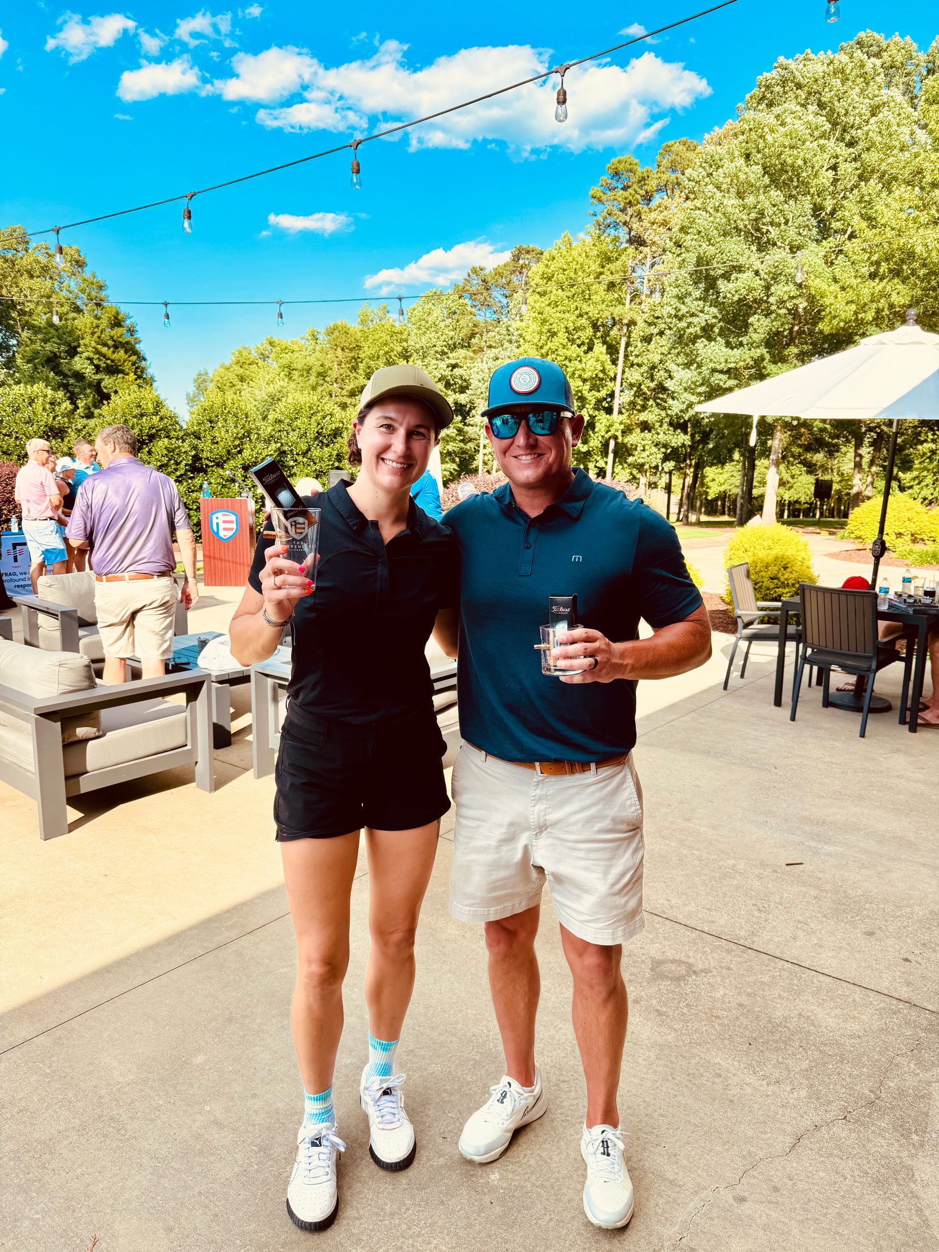 A person in a black outfit and another in a blue polo and white shorts stand together, smiling, on a sunny outdoor patio.