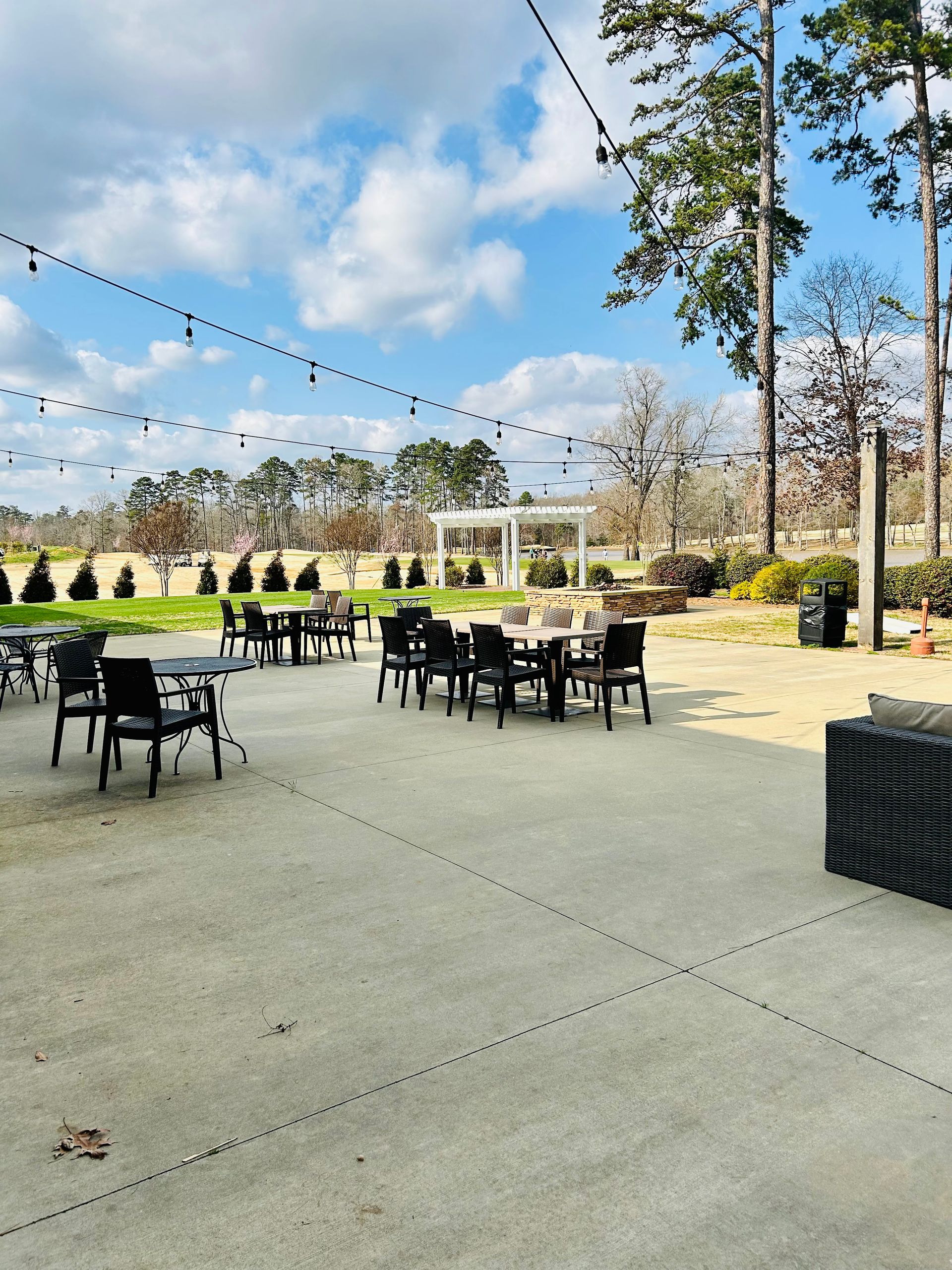 An outdoor patio with scattered tables and chairs under string lights, overlooking a golf course and a white pergola.