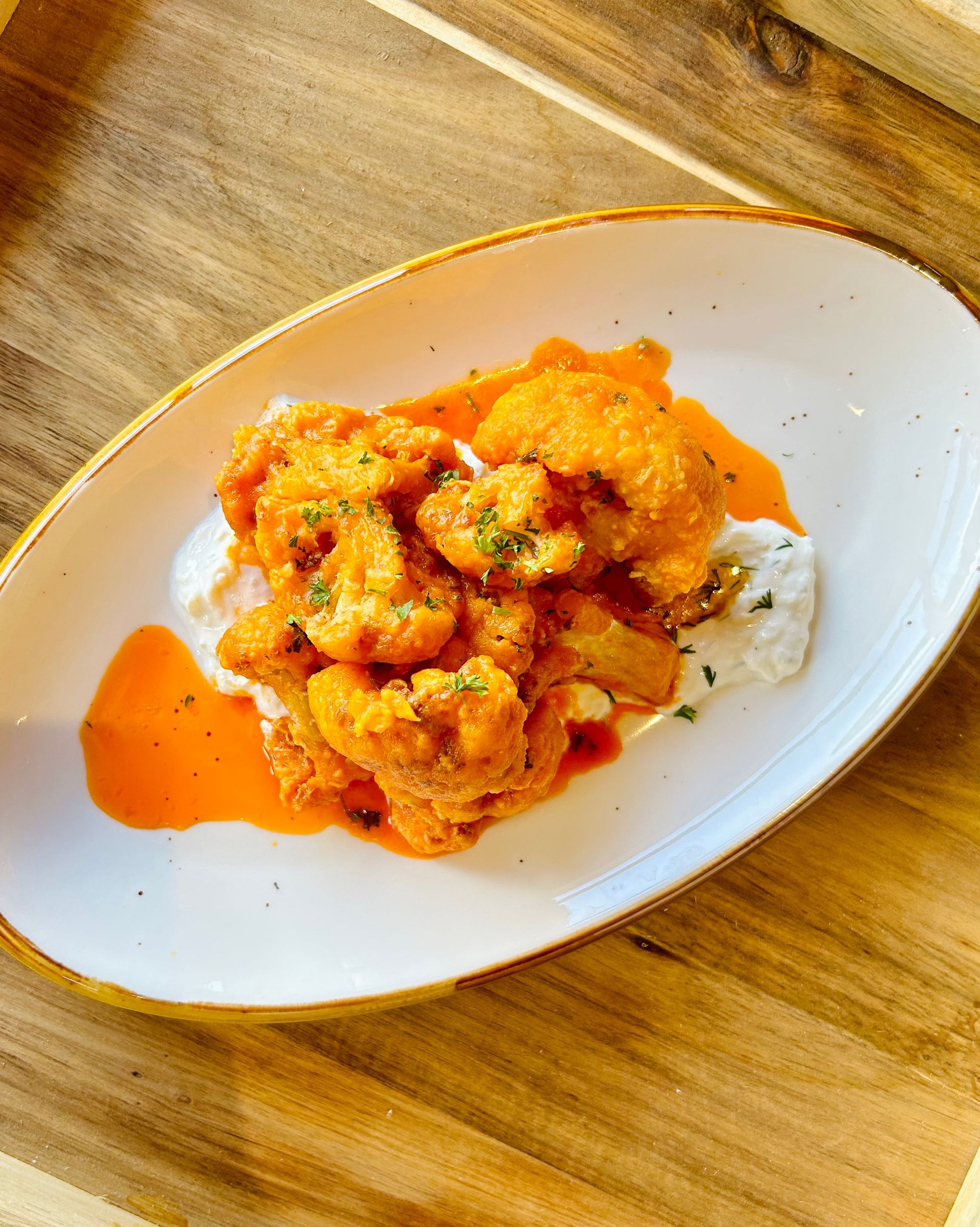Buffalo cauliflower florets tossed in orange sauce, served over a dollop of white sauce on a white plate on wood.
