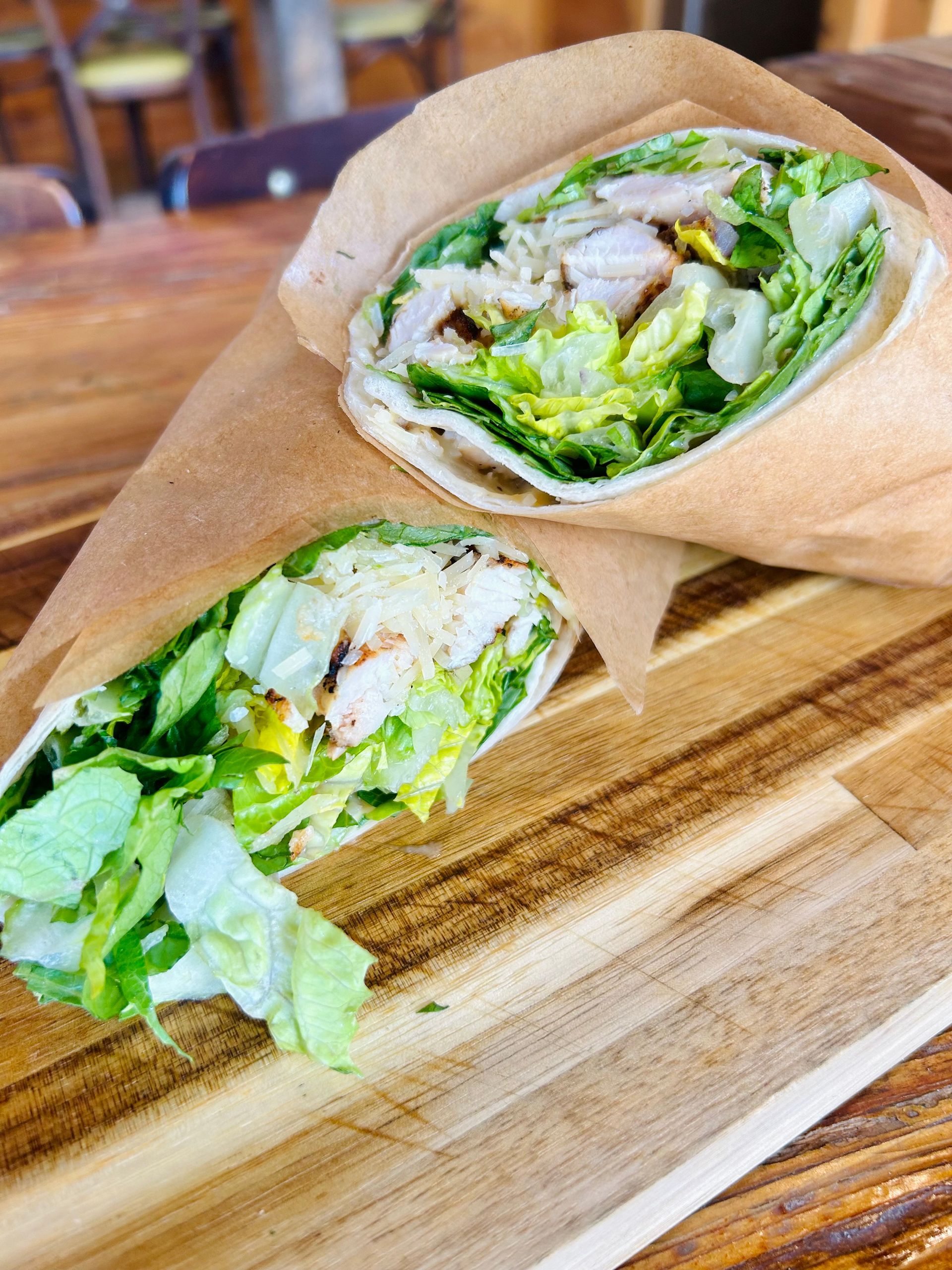 Two chicken Caesar wraps wrapped in brown paper, resting on a wooden cutting board.