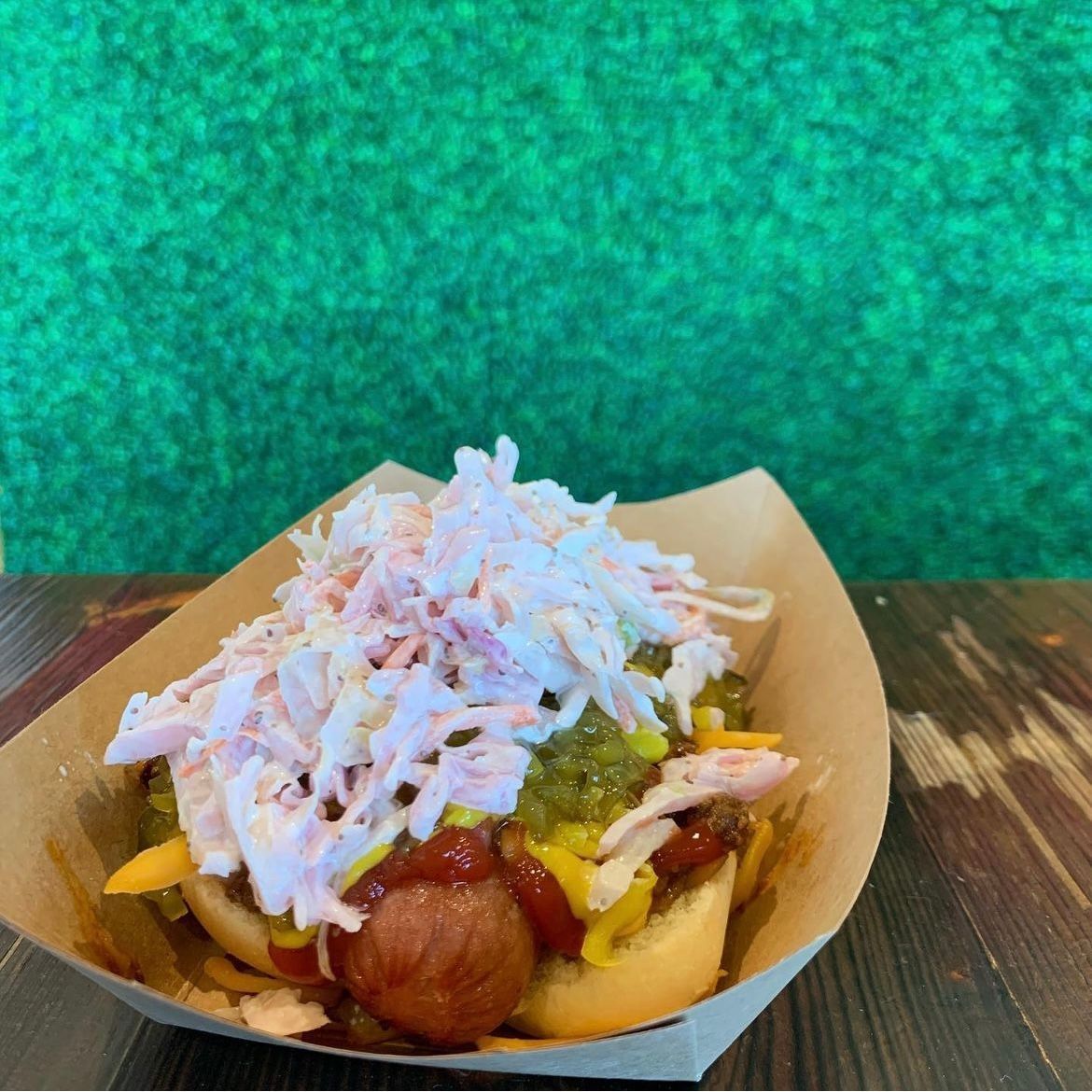 A hot dog in a paper tray topped with relish, shredded cheese, and creamy coleslaw against a textured green background.