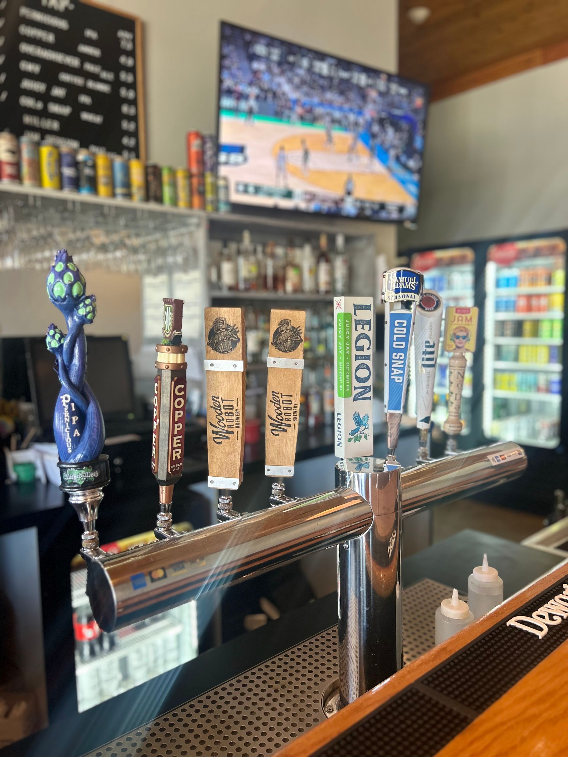 A close-up of a beer tap tower at a bar with a basketball game playing on a television screen in the background.