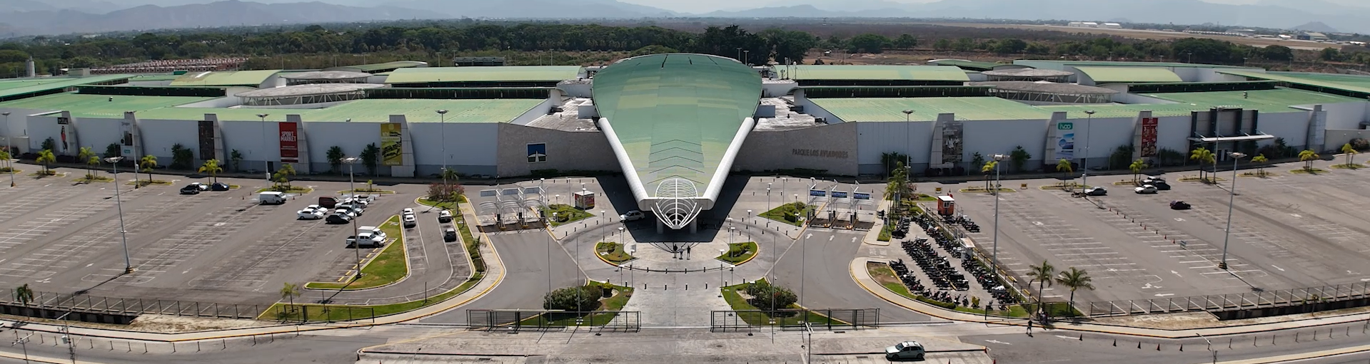 Vista aérea de un moderno edificio con techo de cristal, aparcamientos y zonas ajardinadas a ambos lados.
