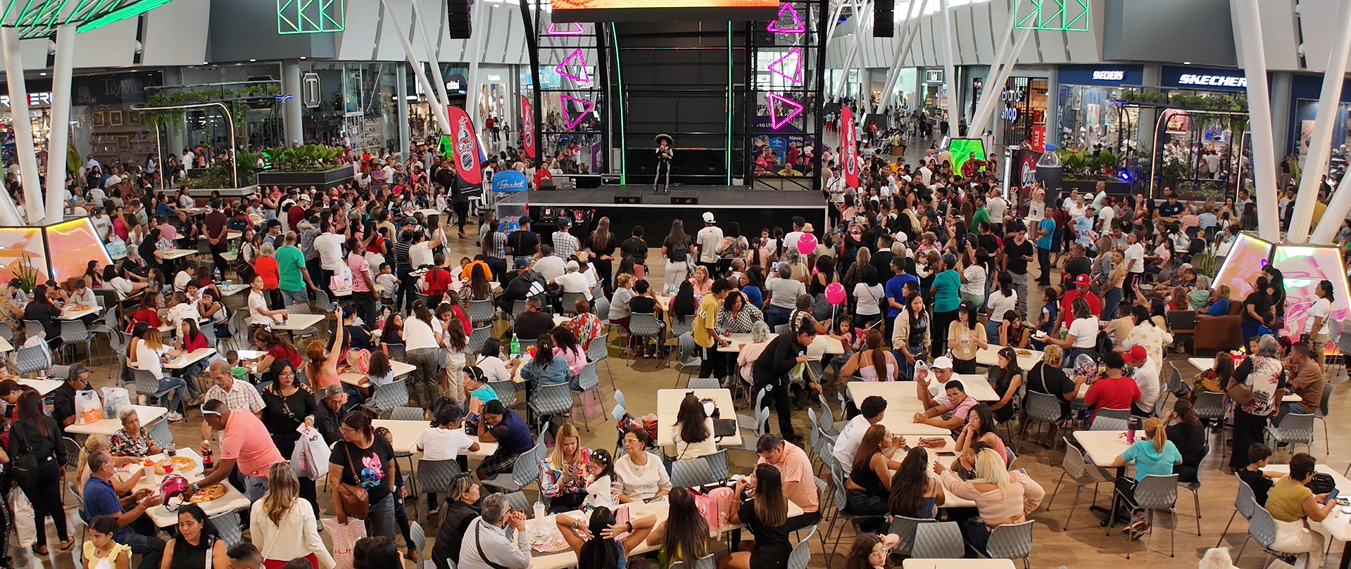 Sala de eventos cubierta y abarrotada de gente, con un gran público frente a un escenario y una pantalla gigante.