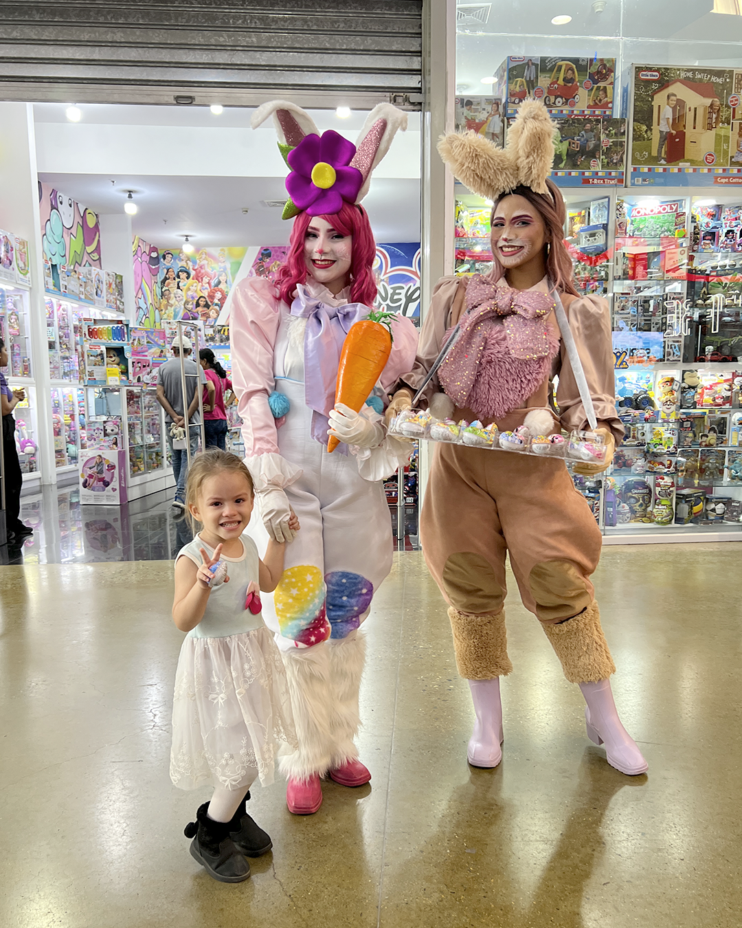 Tres personas con coloridos disfraces de conejos posando en el pasillo de una tienda, con una niña pequeña con un vestido blanco delante.