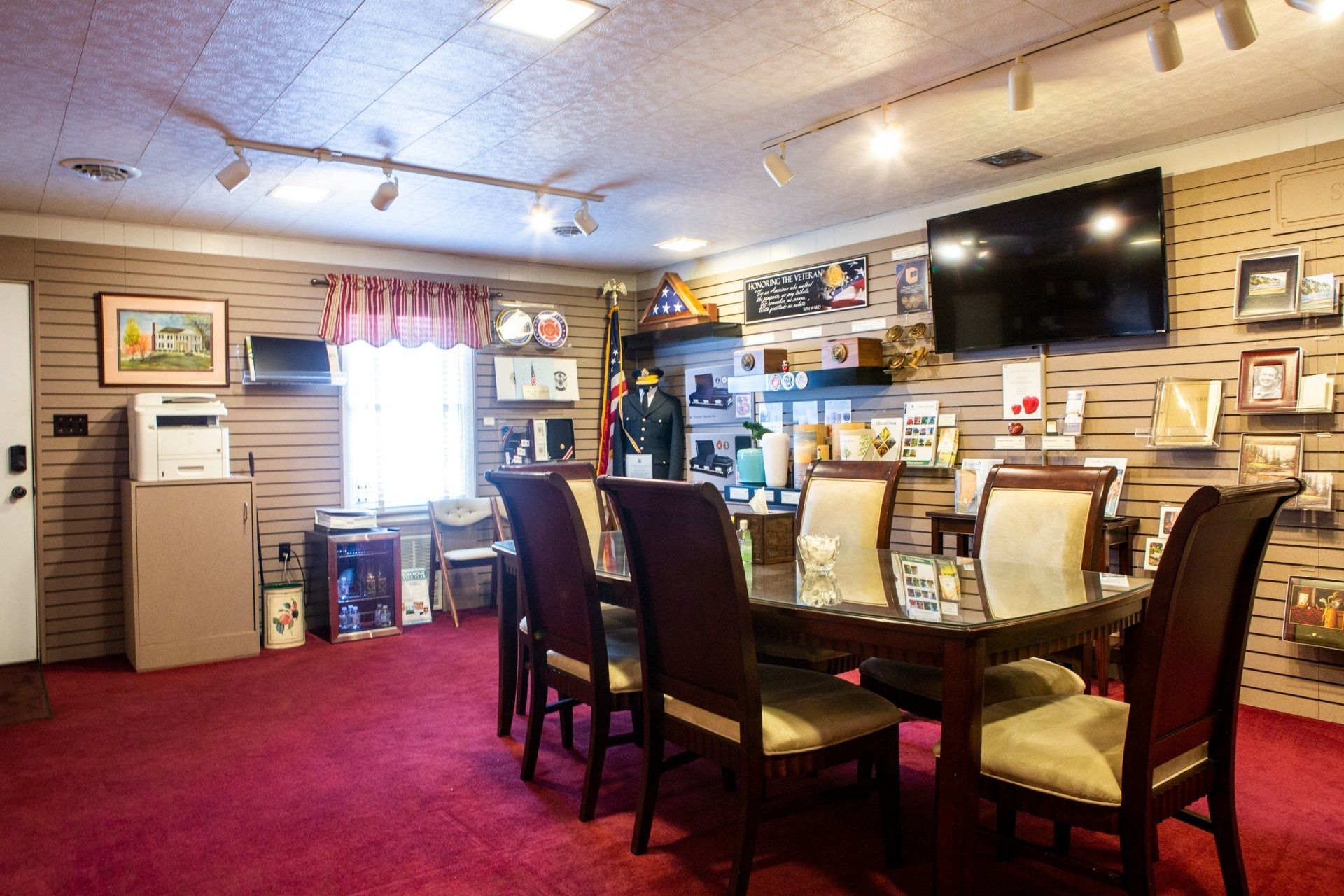 Restaurant dining room with red carpet, tables, chairs, and wall-mounted TV near a window.