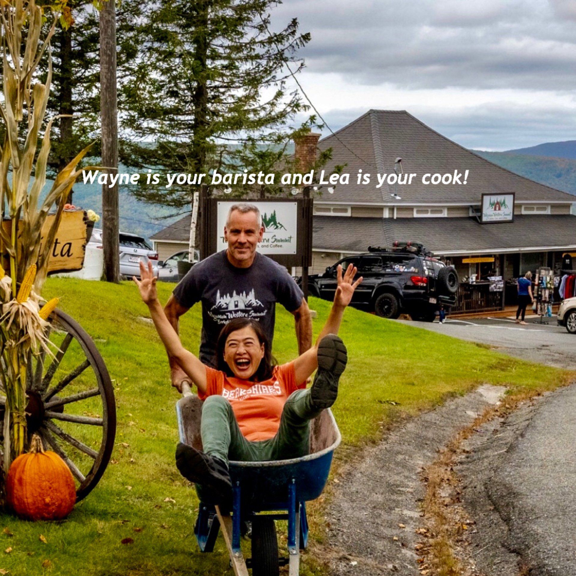 A person pushes another person in a wheelbarrow outdoors in front of a building, with the text:
