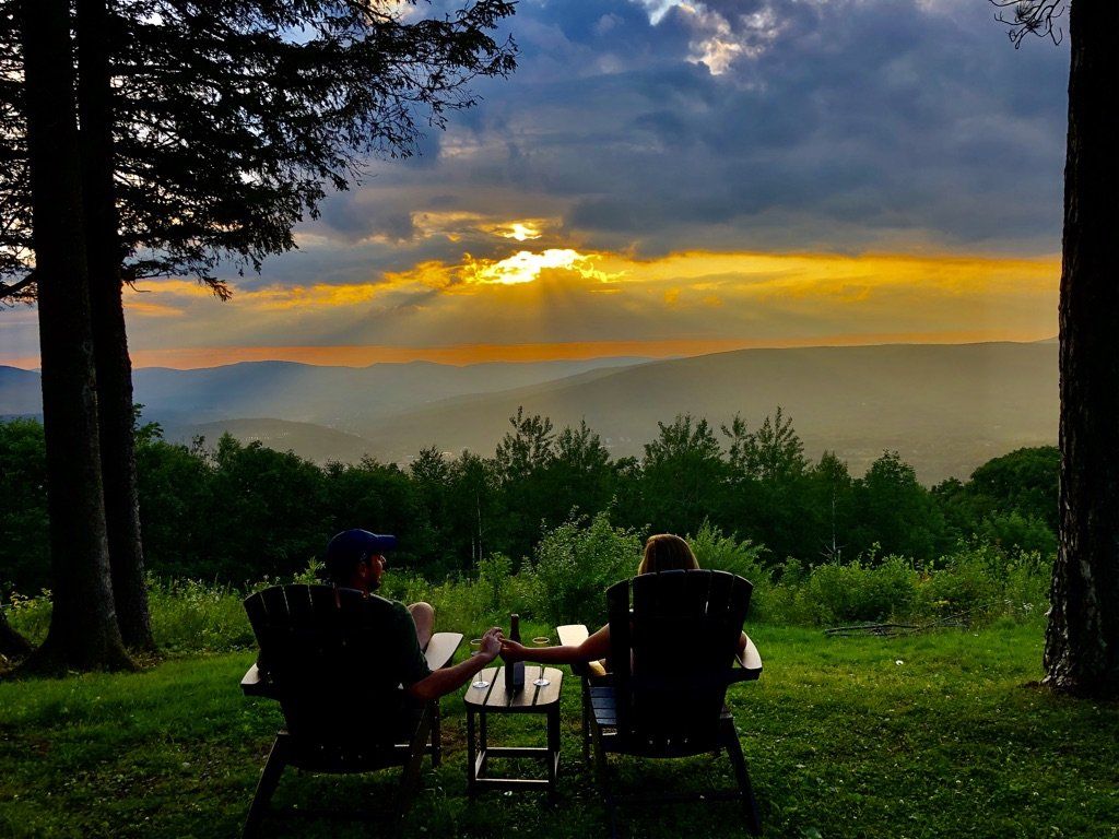 Two people sit in chairs on a grassy hill, holding hands and overlooking a sunset over a distant mountain valley.