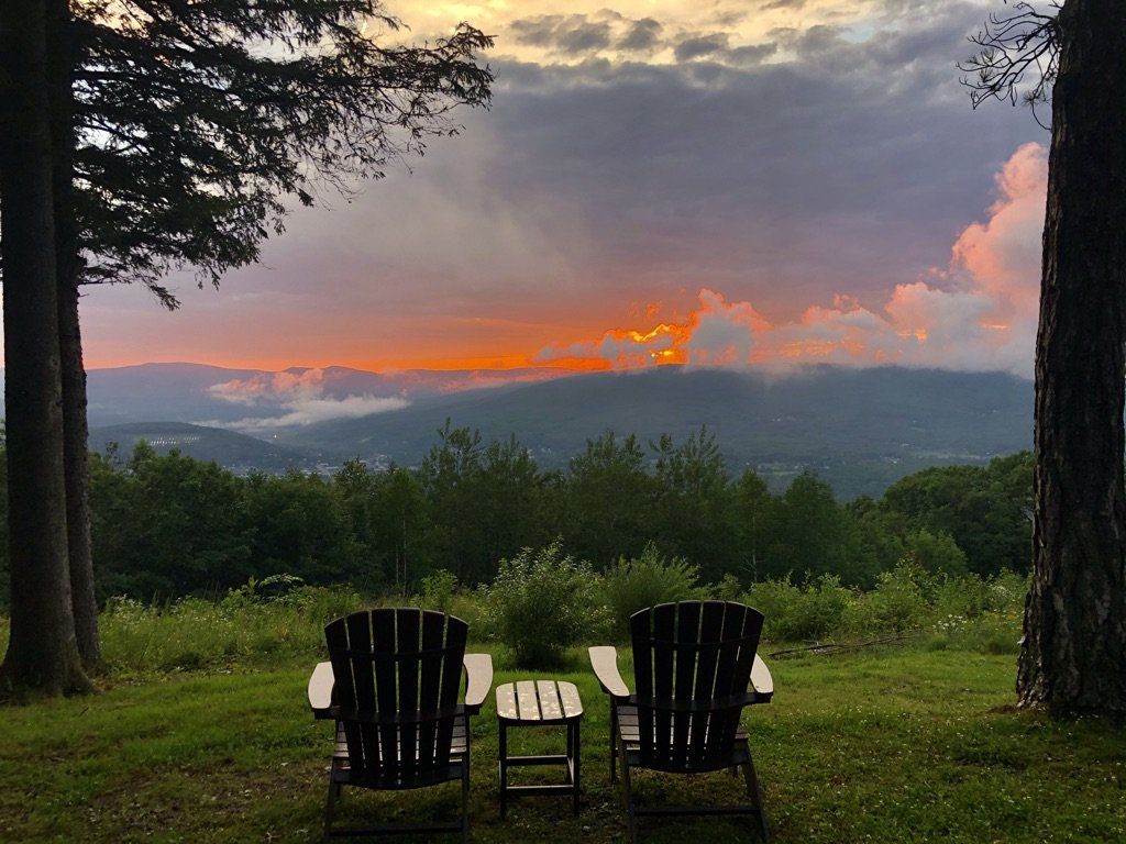 Two Adirondack chairs and a small table face a scenic mountain view during a vibrant sunset with orange and purple skies.