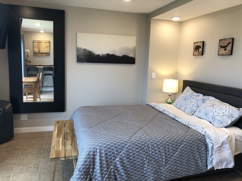 A modern bedroom with a gray patterned bed, a wooden bench, a large black-framed mirror, and two small wall art pieces.