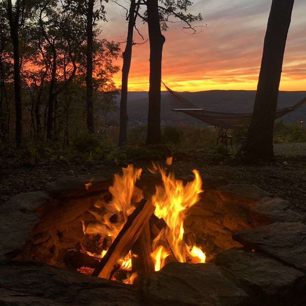 A vibrant sunset glows behind trees over a scenic valley, with a hammock nearby and a bright campfire in the foreground.
