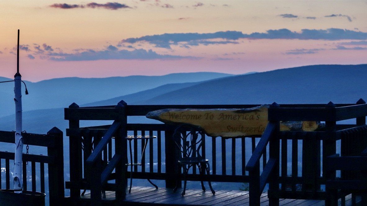 A dark wooden viewing deck overlooking layered mountain ranges under a soft purple and orange sunset sky.